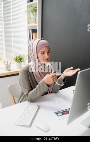 Junge selbstbewusste Lehrerin in Hijab, die vor dem Computermonitor vor dem Online-Publikum spricht, während sie in der häuslichen Umgebung am Schreibtisch sitzt Stockfoto