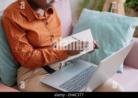 Hände einer jungen afrikanischen Studentin mit Stift, die während des Online-Studiums Vortragsnotizen in einem Copybook macht Stockfoto