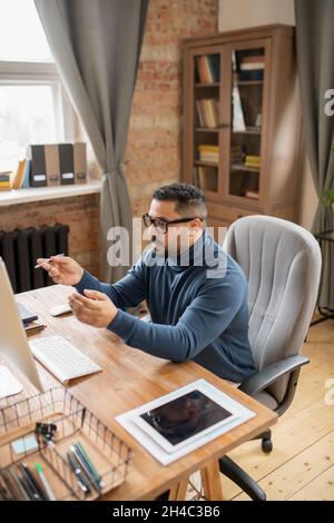 Reifer, selbstbewusster männlicher Lehrer, der vor dem Computermonitor sitzt und dem Online-Publikum Punkte der Lektion erklärt Stockfoto