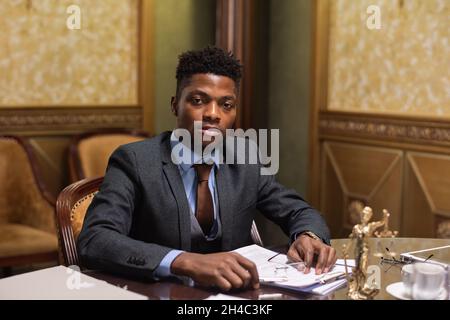 Ein junger seriöser afrikanischer Geschäftsmann oder Anwalt in Anzug schaut Sie an, während er am Tisch im Sitzungssaal sitzt und durch die Papiere schaut Stockfoto
