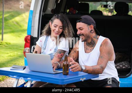 Das junge tätowierte Paar lächelt und benutzt den Laptop in einem Klapptisch neben dem Van. Stockfoto