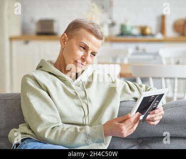 Attraktive, reife Frau, die auf dem Sofa sitzt, das Foto im Rahmen betrachtet und lächelt Stockfoto