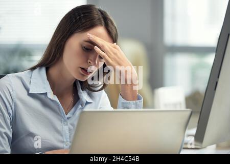 Müde junge kaukasische Geschäftsfrau mit Kopfschmerzen, die die Stirn berühren und einen Laptop benutzen, während sie im Büro arbeitet Stockfoto