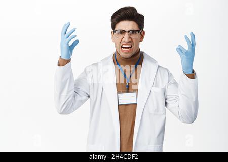 Frustrierter Arzt mit Brille und Gummihandschuhen, der schrie, die Hände schüttelte und wütend aussach und vor weißem Hintergrund verärgert stand Stockfoto