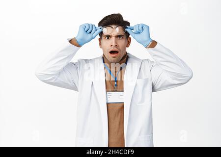 Schockiert und erschrocken zieht der Arzt die Brille aus, keuchend und ungläubig starrend, in medizinischer Robe und Handschuhen auf weißem Hintergrund stehend Stockfoto