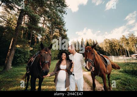 Eine Schwangere in einem Hut mit einem Mann in weißen Kleidern, der mit Pferden in der Natur unterwegs ist. Eine Familie, die auf ein Kind wartet, geht im Wald spazieren. Stockfoto