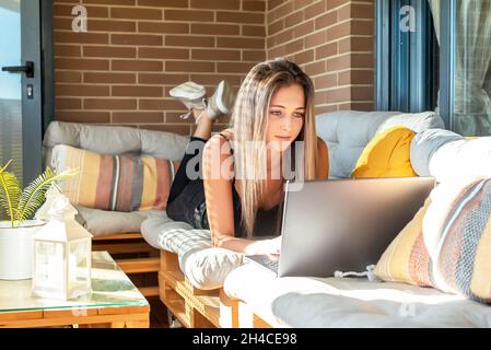 Junge blonde hübsche Frau, die zu Hause mit einem Laptop auf einem Sofa studiert Stockfoto