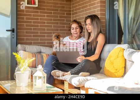 Junge Frau erklärt einer älteren Frau mit einem Welpen, der zu Hause auf dem Sofa sitzt, wie man das Internet nutzt Stockfoto