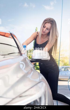 Süße blonde Frau in Jeans und schwarzem T-Shirt trocknet das Auto nach dem Waschen Stockfoto
