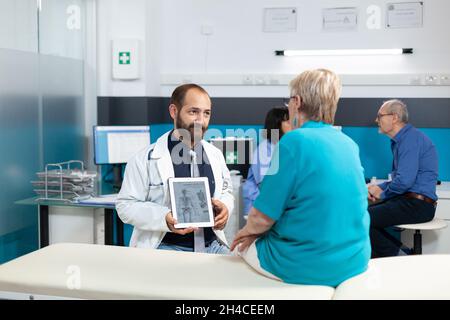 Arzt, der ein menschliches Skelettbild auf einer modernen Tablette zur Osteopathiediagnose zeigt. Medic erklärt älteren Patienten Knochenbrüche am Gerät für orthopädische Versorgung und chiropraktische Genesung. Stockfoto