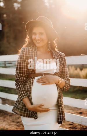 Ein schwanger Mädchen mit einem großen Bauch in einem Hut in der Nähe eines Pferdes Corral in der Natur bei Sonnenuntergang.stilvolle Schwangere in einem braunen Kleid mit Pferden Stockfoto