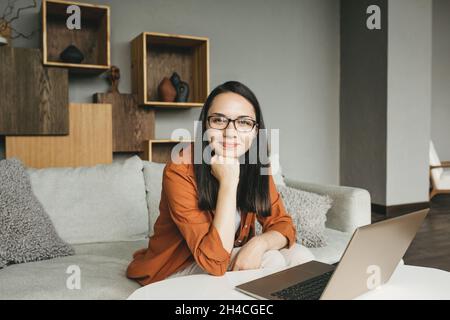 Eine schöne junge europäische Frau in legerer Kleidung, die von zu Hause aus einen Laptop benutzt. Führt einen Videoanruf durch, kommuniziert online mit Kollegen, sendet und antwortet auf E-Mails, entwickelt Software, während Sie zu Hause auf dem Sofa sitzen. Eine kluge Frau arbeitet aus der Ferne Stockfoto