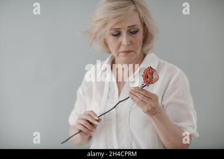 Traurige ältere Frau im weißen Hemd hält eine trockene, alte, tote Rosenblume. Alter und Alter konzeptuell Stockfoto