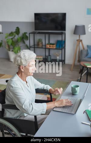 Vertikale Seitenansicht Porträt einer älteren Frau im Rollstuhl, die zu Hause einen Laptop benutzt Stockfoto