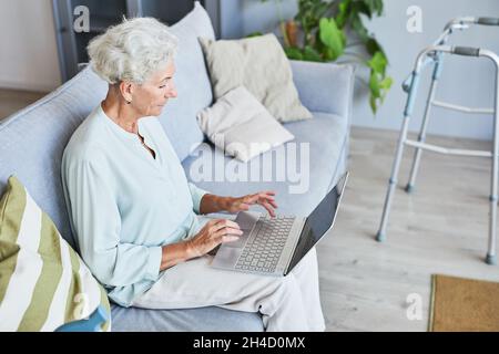 Seitenansicht Porträt einer weißhaarigen älteren Frau, die einen Laptop benutzt, während sie sich auf dem Sofa zu Hause entspannt und Platz kopiert Stockfoto