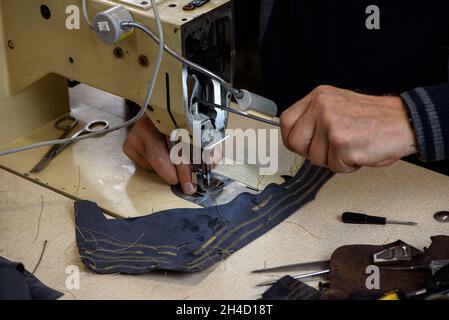 Hände des Reparaturers, der Probleme mit professioneller Industrienähmaschine in der Schuster-Werkstatt behebt. Prüfung der Maschinenheftung. Selektiver Fokus Stockfoto