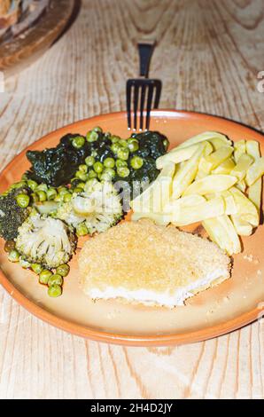 schnitzel mit gedünstetem Gemüse und Pommes in einem Gericht auf einer Holzplatte Stockfoto