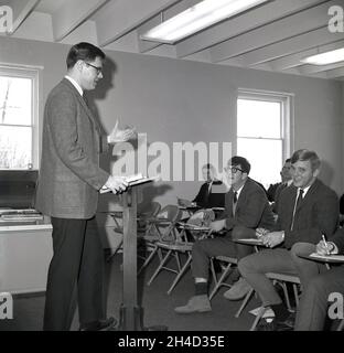 In den 1960er Jahren sitzen historische, gut verkleidete ältere männliche Studenten an einer Geschäftshochschule in einem Raum auf Metallstühlen mit kleinen Schreibtischen und hören einem männlichen Lehrer zu, der an einem hölzernen Rednerpult, Monroe College, Middletown, Virginia, USA, steht. Stockfoto