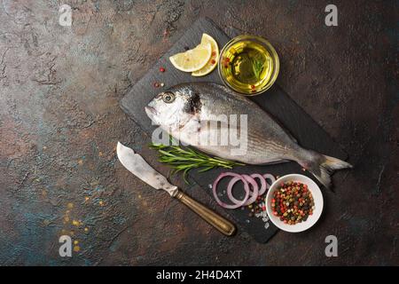 Draufsicht auf frischen Fisch dorado oder Goldbrasse mit Zitrone, Olivenöl, Zwiebeln und Gewürzen wie Pfeffer, Rosmarin, Salz auf schwarzem Schieferbrett und brauner Konz Stockfoto