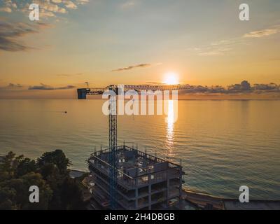 Drohnenansicht eines Turmkrans und eines Gebäudes im Bau vor dem Hintergrund eines fantastischen Sonnenuntergangs an der Küste. Stockfoto