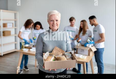 Glückliche ältere Frau, die einen Karton mit Spenden, Essen und einem Lächeln auf die Kamera hielt, während eine Gruppe Freiwilliger Boxen packte Stockfoto