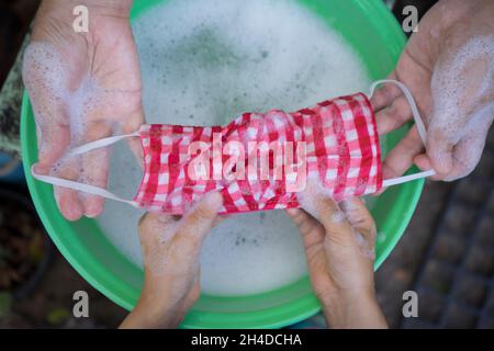 Mama und Kinder Hand Waschen einer Maske mit Waschmittel in Wasser gelöst. Das Waschen der Masken desinfiziert und spart Geld, um wieder verwendet zu werden. Stockfoto