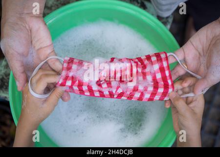 Mama und Kinder Hand Waschen einer Maske mit Waschmittel in Wasser gelöst. Das Waschen der Masken desinfiziert und spart Geld, um wieder verwendet zu werden. Stockfoto