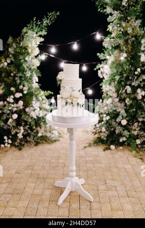 Weiße Hochzeitstorte mit weißen Blumen verziert. Der Kuchen steht vor dem Hintergrund des Abendbogens. Stockfoto