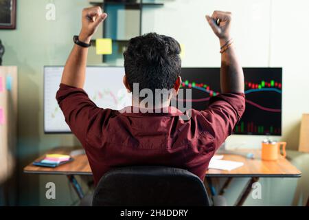 Rückansicht des erfolgreichen Trader, der sich für den Gewinn von Aktien durch das Betrachten von Charts oder Grafiken am Computer im Büro feiert - Konzept des ausgegebenen Investors Stockfoto