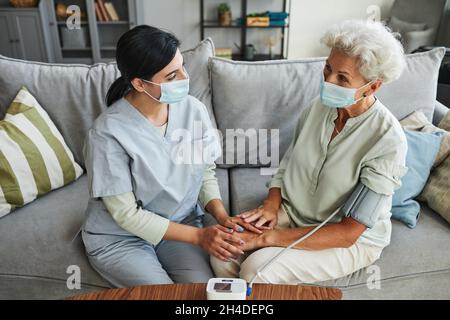 Hochwinkelporträt einer Krankenschwester, die den Blutdruck einer älteren Frau im Altersheim misst, beide mit Masken Stockfoto