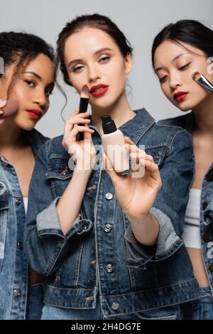 Gesichtsfundament in der Hand einer verschwommenen Frau in der Nähe von interracial Freunden mit kosmetischen Bürsten auf grau isoliert Stockfoto