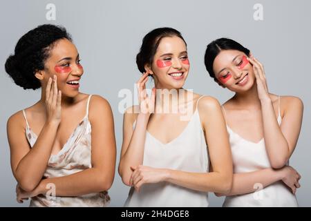 Hübsche interracial Frauen mit Augenflecken berühren Gesichter isoliert auf grau Stockfoto