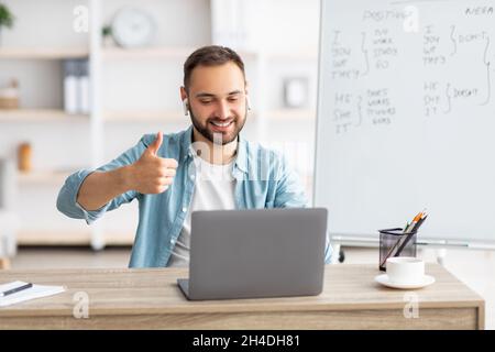 Glücklicher männlicher Lehrer, der mit einem Laptop Englisch im Internet unterrichtet, den Daumen hochgestikelt und eine Fremdsprachenschule empfohlen hat Stockfoto