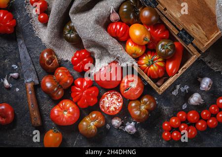 Stillleben in der Küche mit Retro-Objekten, Draufsicht - mehrfarbige Tomaten in verschiedenen Größen und Typen, Knoblauch, Messer, Schachtel, Sackleinen. Das Konzept des Kochs Stockfoto