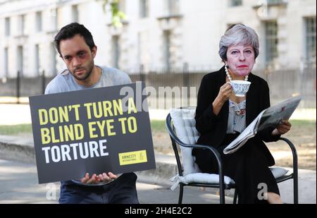 Eine Aktivistin, die eine Maske von Premierministerin Theresa May trägt, inszeniert eine Fotokonferenz vor der Downing Street, während sie eine Tasse Tee trinkt und die Zeitung liest, während sie drei Folteropfer in einem Stunt ignoriert, was einen Aufruf von Amnesty International zur Untersuchung der britischen Beteiligung an Folter hervorhebt. Bild datiert: Montag, 6. August 2018 Stockfoto