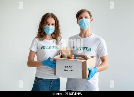 Wohltätigkeitszentrum. Millennial-Freiwillige halten eine Schachtel mit Lebensmittelspenden mit Produkten und Konserven und tragen medizinische Masken Stockfoto