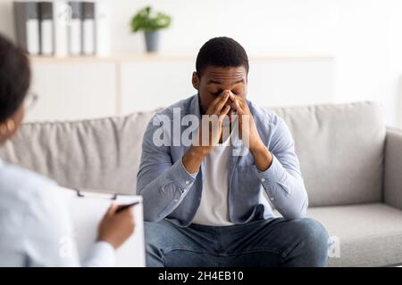 Gestresster schwarzer Kerl, der Sitzung im Büro des Psychologen hat und professionelle Hilfe bei der Lösung persönlicher Probleme erhält Stockfoto