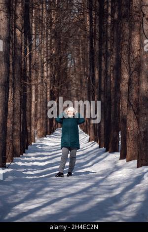 Junge Frau steht auf der Straße zwischen Bäumen. Sie hält weißen Hut mit ihren Händen, starker Wind weht und es ist kalt im Wald Stockfoto