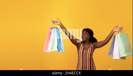 Aufgeregt junge schwarze Frau, die die Arme hochhebt, helle Einkaufstaschen hält und auf leeren Raum für Ihre Werbung schaut Stockfoto
