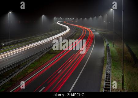 Weiße Scheinwerfer und rote Rücklichter an einem nebligen Abend auf der Autobahn A44 in der Nähe des Dorfes Abbenes in den Niederlanden. Langzeitbelichtung. Stockfoto