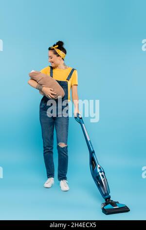 Fröhliche Frau in Denim-Overalls, die Babypuppe hält, während sie auf Blau saugt Stockfoto