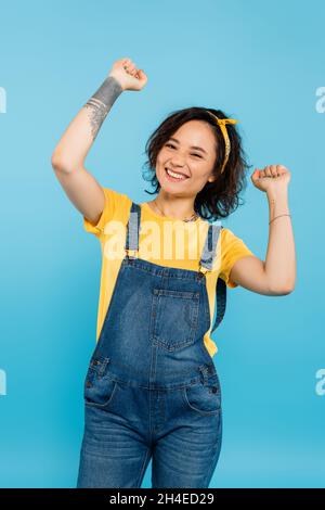 Aufgeregte Frau in Denim-Overalls, die eine Siegesture zeigt, während sie die Kamera isoliert auf Blau betrachtet Stockfoto