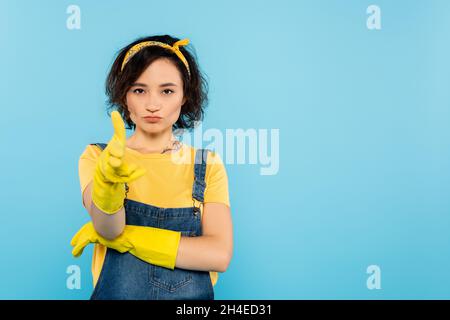 Frau in Denim-Overalls und Gummihandschuhen, die auf blau isolierte Gefussture zeigen Stockfoto