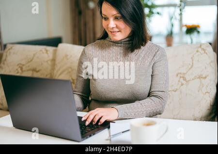 Frau arbeitet auf einem Laptop aus der Ferne auf dem Hintergrund des Innenraums des Wohnzimmers. Geschäftsfrau 30-40 Jahre alt sitzt auf der Couch zu Hause und arbeitet o Stockfoto