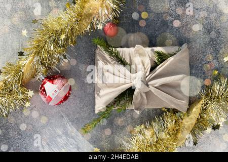 Geschenk im Furoshiki-Stil für Weihnachten oder Neujahr auf grauem Hintergrund mit Bokeh. Flach mit Kugeln und Lametta legen. Stockfoto
