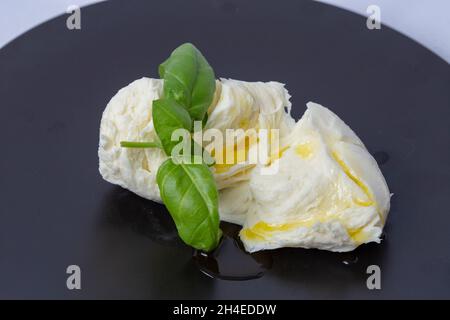 Zerdrückte Mozzarella-Kugel, mit Olivenöl und Basilikumblättern, auf einem schwarzen Teller Stockfoto