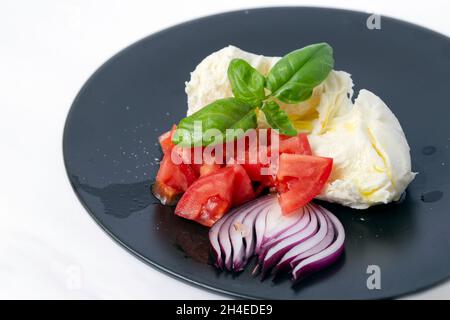 Mozzarella-Käse mit Tomaten, Basilikum, Zwiebeln und Olivenöl, auf einem schwarzen Teller. Stockfoto