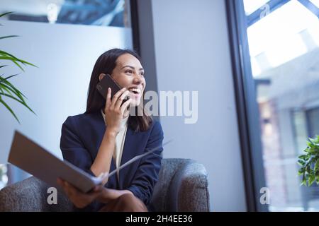 Biracial Geschäftsfrau lächeln, halten Dokumente und sprechen auf dem Smartphone in modernen Büro Stockfoto