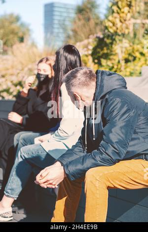 Neu ist normal, Covid-19, soziales Distanzkonzept. Eine Gruppe von Freunden mit schützenden Gesichtsmasken sitzt und spricht während einer Pandemie auf der Bank. Helle Viv Stockfoto