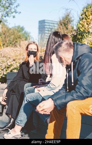 Neu ist normal, Covid-19, soziales Distanzkonzept. Eine Gruppe von Freunden mit schützenden Gesichtsmasken sitzt und spricht während einer Pandemie auf der Bank. Helle Viv Stockfoto